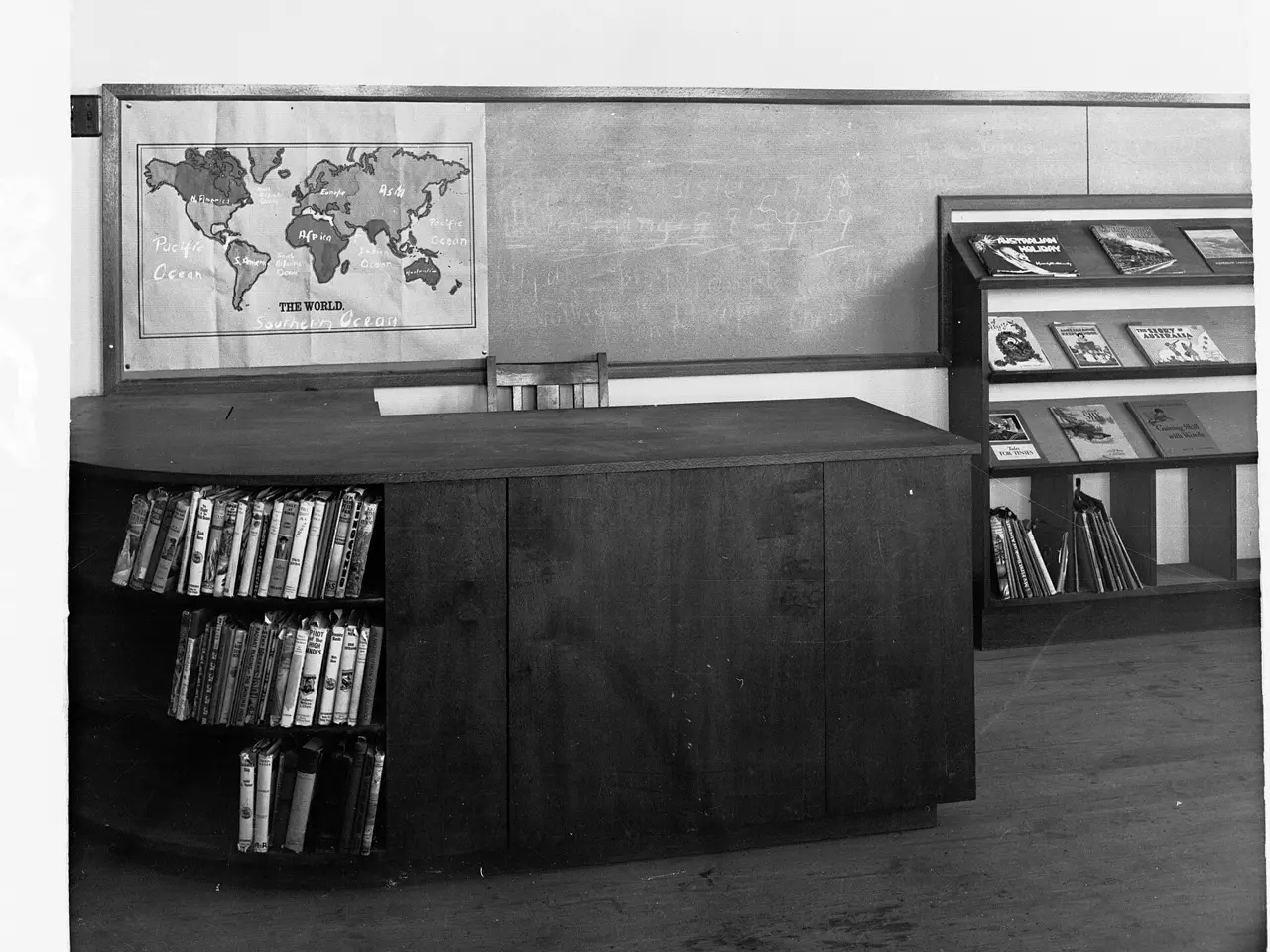 A black and white photograph of an empty school library, with a world map pinned above a desk and shelves of books.